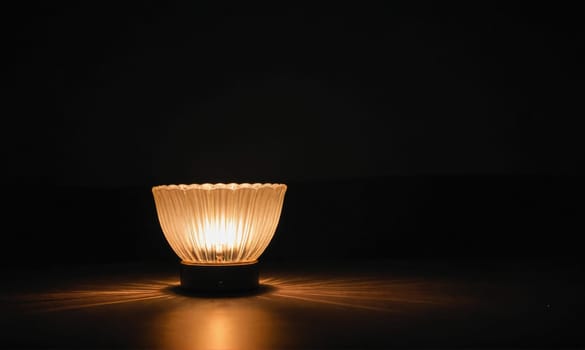 A small lamp with a glass shade illuminates a dark room, casting a warm, golden light on the table surface.