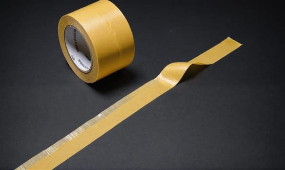 A roll of brown tape is laying on a black surface. A long piece of tape is unrolled and laying next to the roll.