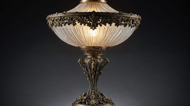 A large, ornate lamp with a clear glass shade rests on a dark surface. The lamp is made of brass and has a decorative base with a scrolled design.