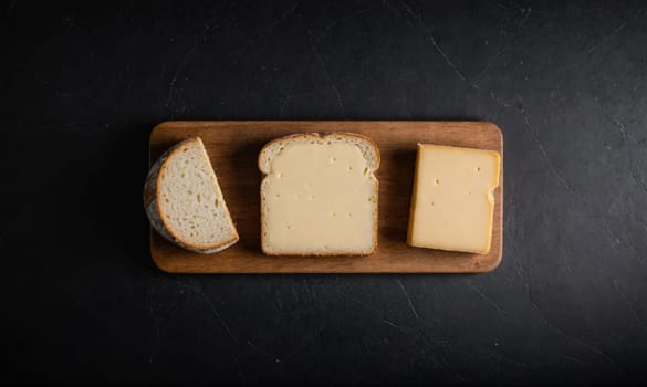 A wooden cutting board sits on a dark background with three slices of white bread and a block of cheese arranged on top.
