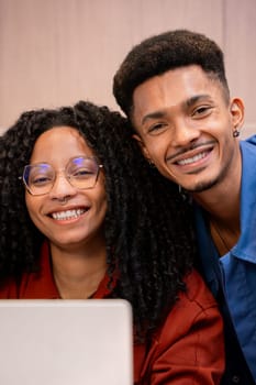 Two cheerful friends, one with curly hair and glasses, share a moment while working on a laptop. Both are smiling and looking at the camera, creating a warm and collaborative atmosphere.
