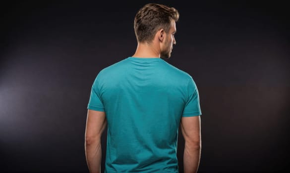 A man is standing in front of a dark background, looking over his shoulder. He is wearing a plain, light blue t-shirt. His arms are at his sides, and his hands are in his pockets.