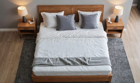 A wooden bed frame with a white duvet and grey pillows sits in a bedroom. The bed is on a grey rug and the floor is made of wood. There are two nightstands with lamps on either side of the bed.
