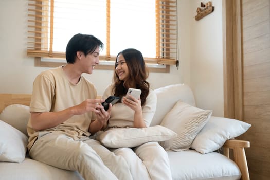 Happy millennial Asian couple using a smart phone together on sofa at home.