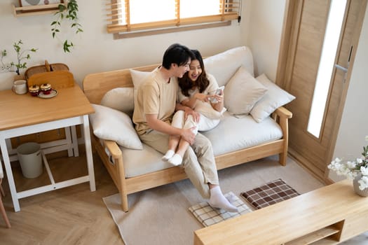 Happy millennial Asian couple using a smart phone together on sofa at home.