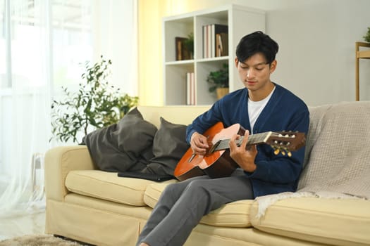 Young adult man relaxing at home playing acoustic guitar on the couch.