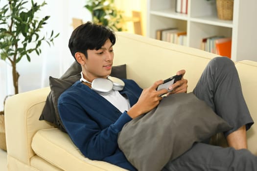Relaxed young asian man enjoying mobile gaming on the couch.