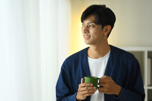 Relaxed young adult man holding a coffee cup gazing outside with a smile.