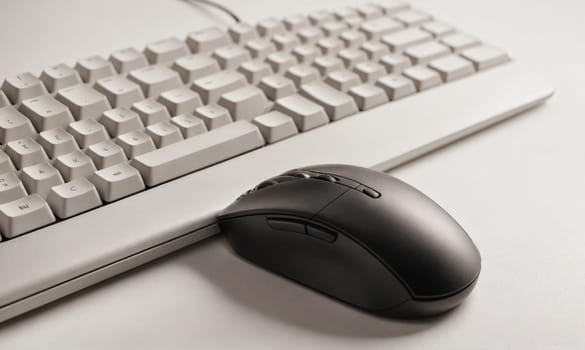 A white keyboard lies on a white desk, with a black mouse sitting on the desk below it.