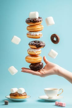 Hand is holding a cup catching flying donuts, chocolate bars and marshmallows on a blue background. This image evokes the concept of sweet breakfast