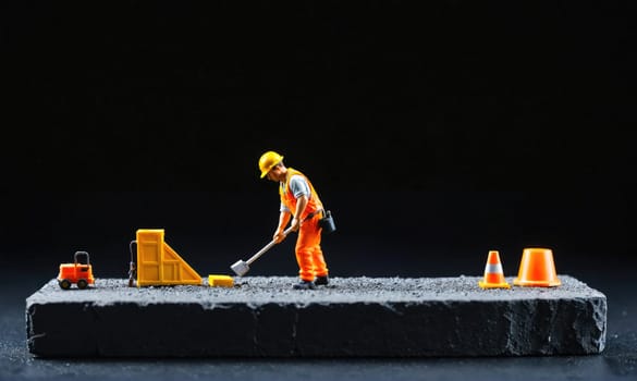 A miniature construction worker wearing an orange jumpsuit and a yellow hard hat is using a shovel to move dirt in a small-scale construction scene.