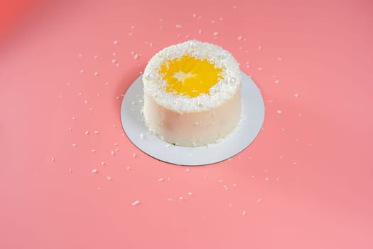 Delicious white cake decorated with orange slice and coconut flakes standing on pink background with scattered coconut flakes