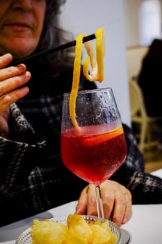 Woman adding a lemon zest to a red cocktail in a wine glass, with a bowl of chips on the table