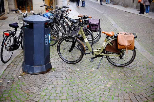 Cremona, Lombardy, Italy - November 7th 2024 Parked bicycles and a public trash can rest on a cobblestone street with pedestrians walking in the background