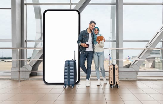 A cheerful muslim couple stands together by huge phone with blank screen at the airport, with luggage nearby, while they engage with their devices
