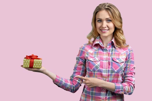 Attractive young blond woman is pointing at gift box. Isolated on pink background.