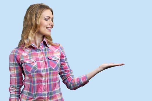 Smiling young blond woman is presenting copy space with one hand. Isolated on pastel blue background.