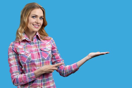 Smiling young blond woman holding palm up and pointing with her finger. Isolated on vivid blue background.