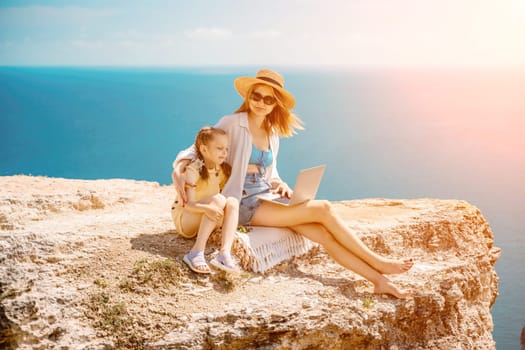 Freelance woman with her daughter working on a laptop by the sea, typing on the keyboard, enjoying the beautiful view, highlighting the idea of remote work