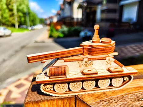 Wooden toy tank with Russian inscription t-34 and city or village houses on sunny day. War between Russia, Ukraine. Concept of defense, attack on civilian population by military equipment