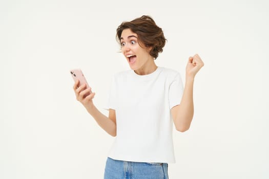 Portrait of excited brunette woman with smartphone, looking at mobile phone, screams surprised from amazement and joy, winning online, standing over white background.