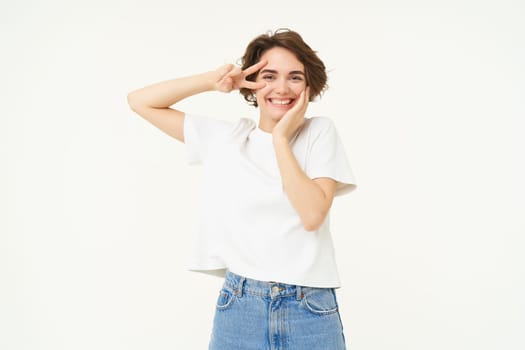 Portrait of happy and carefree woman, shows peace, v-sign gesture, having fun, posing for photo over white background.