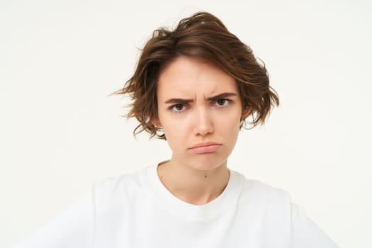 Portrait of frowning, grumpy young woman, looks upset and offended, disappointed by something, standing over white background. Copy space