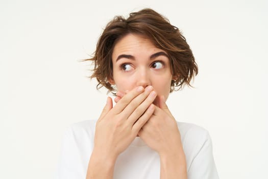 Oops. Girl looks around, said something wrong, shuts her mouth, covers lips with hands and makes side eye, stands over white background.