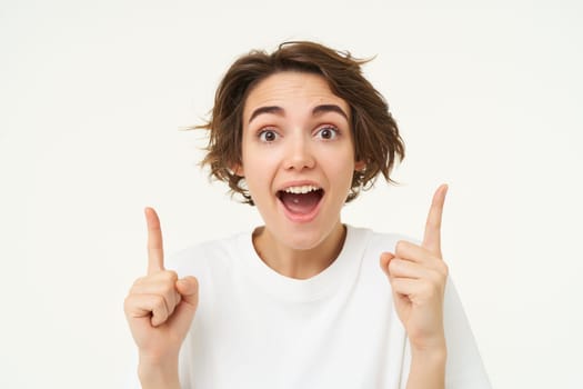 Portrait of happy, beautiful young woman pointing fingers up, showing advertisement or product on top of copy space, standing against white background.