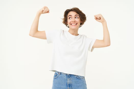 Young happy girl feeling strong, woman shows biceps, flexing muscles on arms and smiling, proves her strength, stands over white background.