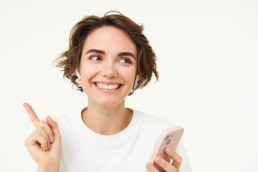 Portrait of brunette woman listening music in headphones, pointing left at banner, holding mobile phone, using streaming app, isolated over white background.