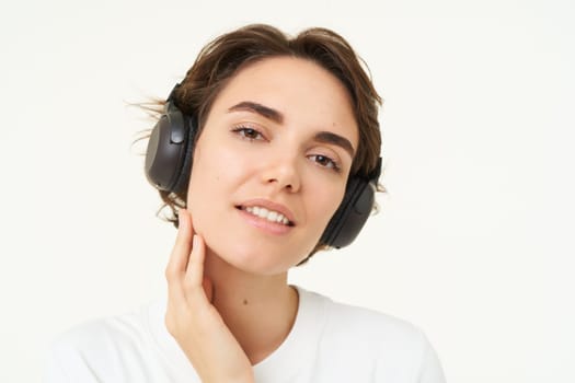 Portrait of young candid woman in wireless headphones, smiling, listening music in earphones, standing over white background.