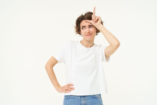 Portrait of young woman making fun of someone, showing loser sign on forehead and looking arrogant, standing over white background.