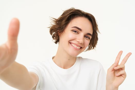 Close up of smiling brunette woman makes selfie, taking photo for her social media, holds mobile phone with extended hand, standing over white background.