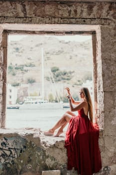 Side view a Young beautiful sensual woman in a red long dress posing on a volcanic rock high above the sea during sunset. Girl on the nature on overcast sky background. Fashion photo