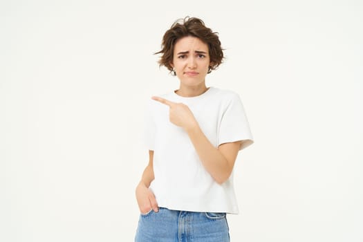 Portrait of sceptical young woman, pointing finger left and looking with doubt at camera, dislike smth, standing over white studio background.