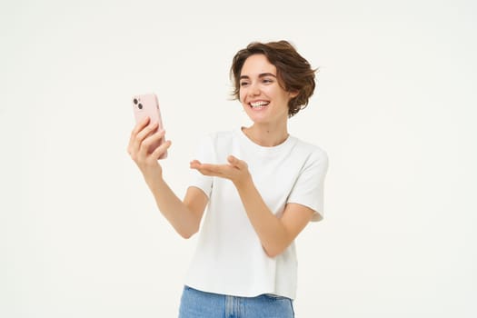 Portrait of girl talking on mobile phone, video chats with friends on smartphone, standing over white studio background.