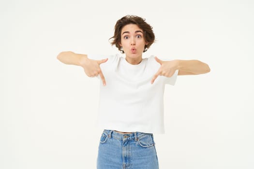 Portrait of amazed girl, pointing fingers down, showing something and looking surprised, standing over white studio background.