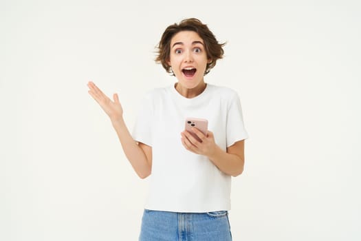 Portrait of brunette girl expresses amazement and surprise, holds smartphone, reacts to amazing advertisement on mobile phone, standing over white background.