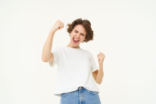 Cheerful brunette woman winning, triumphing, celebrating victory, achieves goal, stands over white background.