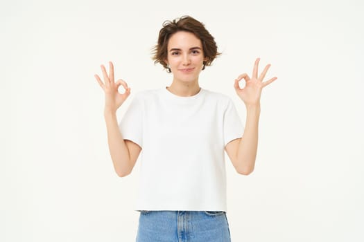 Portrait of young brunette woman, look confident, shows okay, ok sign, approves something, recommending, give positive feedback, stands over white background.