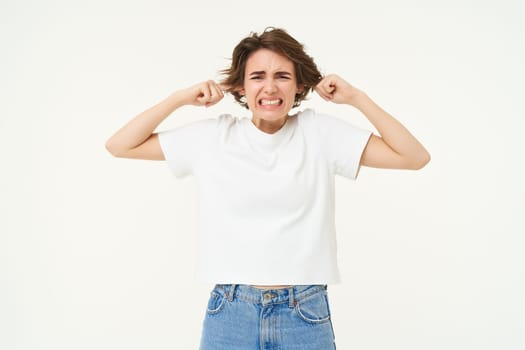 Portrait of upset woman shuts her ears, feeling discomfort from loud noise, standing over white background. Copy space