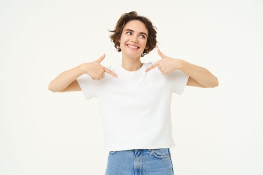 Image of brunette female model, pointing at herself and smiling, isolated against white studio background.