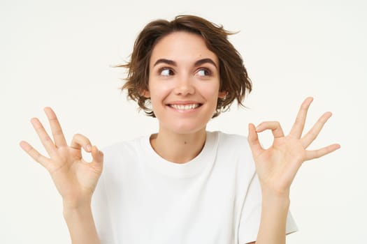 Image of excited brunette woman, smiles, shows okay, ok gesture, looks amused and happy, stands over white studio background.