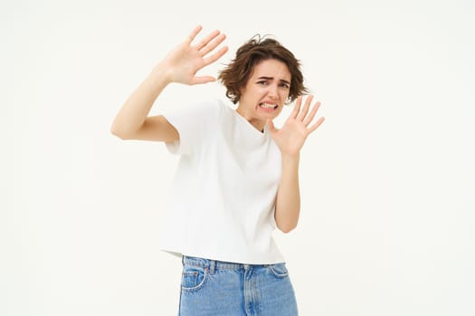 Portrait of woman raising hands in defense and blocking face from something, standing over white background. People and emotions concept