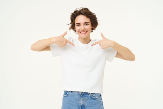 Image of confident, good-looking young woman, smiling and pointing at herself, standing over white studio background. Copy space