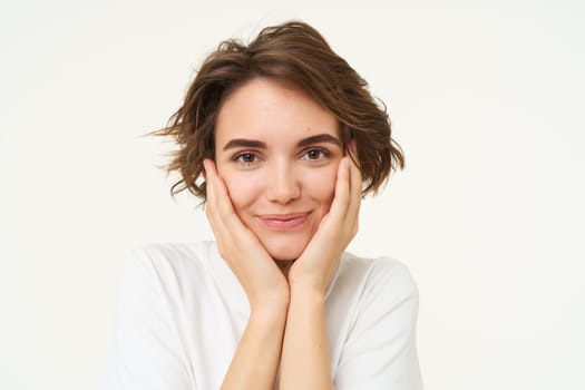 People and emotions concept. Young woman touches her face, smiling, pleased with her face condition after skincare treatment, stands over white background.