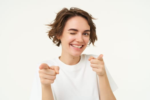 Close up portrait of cute smiling woman, pointing fingers at camera, choosing, inviting you, posing against white studio background.