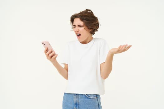 Portrait of frustrated girl with smartphone, woman looking at telephone and reacting shocked at mobile phone, shrugging shoulders, standing over white background.