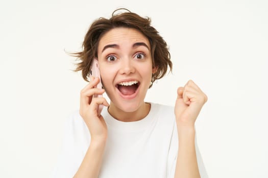 Portrait of amazed brunette girl looks surprised while talks on mobile phone, answers a call, amazed by big news, gasping shocked, standing over white background.